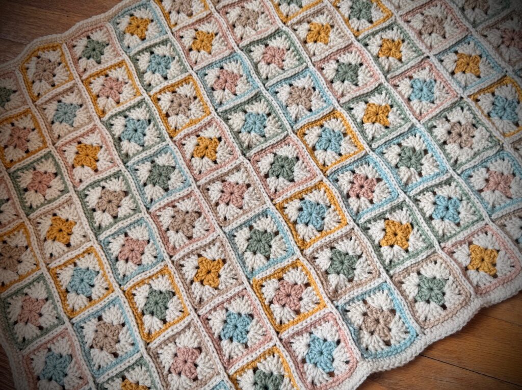 Finished crochet granny square blanket in soft pastel colors with cream joining and border, laid flat to show full layout and texture.