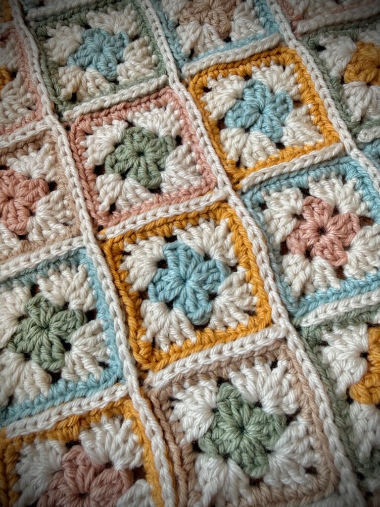 Close-up of a crochet granny square blanket with soft pastel colors and cream joining, highlighting textured stitches and color contrast between squares.